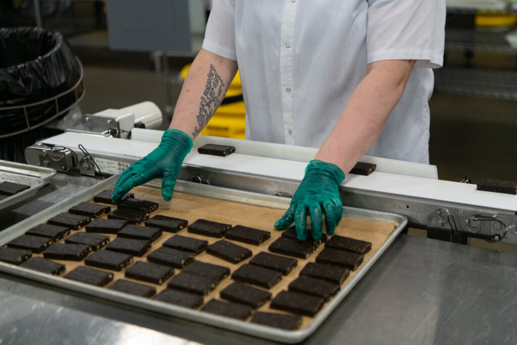 Production worker in gloves arranging rows of chocolate-coated bars on a parchment-lined tray in a food manufacturing facility