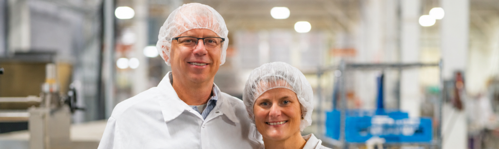 Glenn and Liz Ward standing together in white smocks and hairnets on the floor of a food manufacturing facility with stainless steel production equipment behind them.