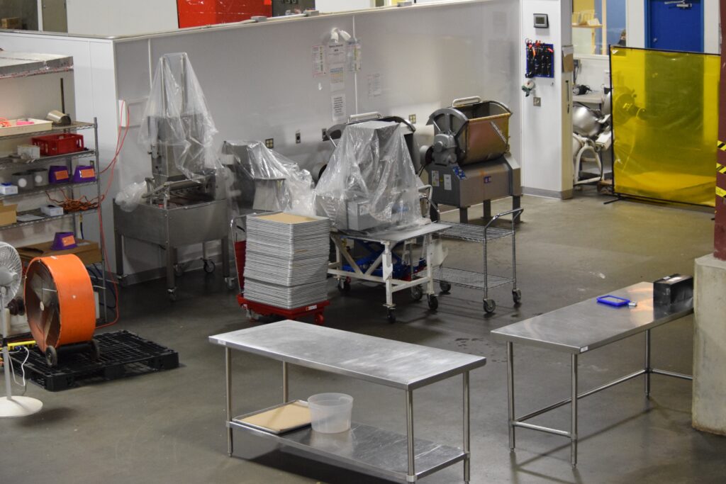 Food processing room with covered mixers, stainless prep tables, and a strip curtain, cleaned and ready for the next shift