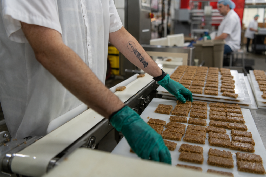 Gloved production worker arranging rows of bars on a conveyor belt in a food manufacturing facility