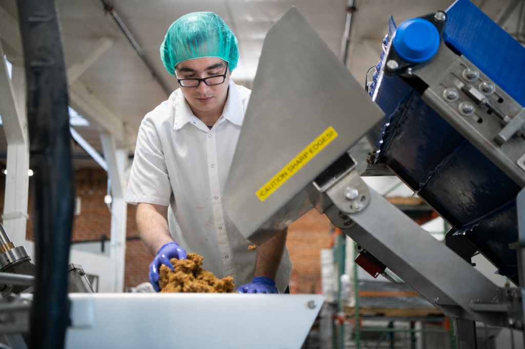 Food production worker in a hairnet and gloves loading granola mixture into a stainless steel hopper on a manufacturing line