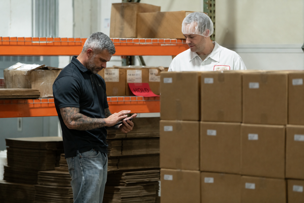 Two food production workers reviewing inventory data on a tablet among stacked boxes on warehouse shelving