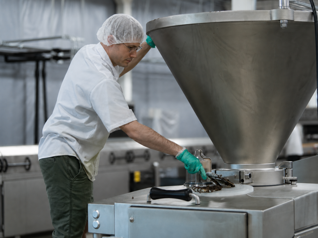 Food production worker in a hairnet operating a large stainless steel funnel hopper on a filling machine