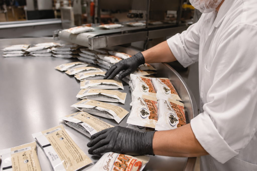 A worker in a white coat, hairnet, and black gloves pulls individually wrapped gluten-free BumbleBar snack bars off a stainless steel rotary accumulation table to pack them in a food production facility.